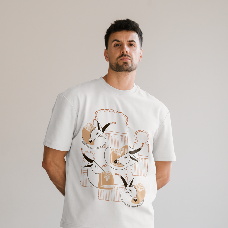 A man wearing a white crewneck t-shirt with a graphic print of illustrated Khillar cow breed of maharashtra with wada style architecture around, set against a plain light gray background.