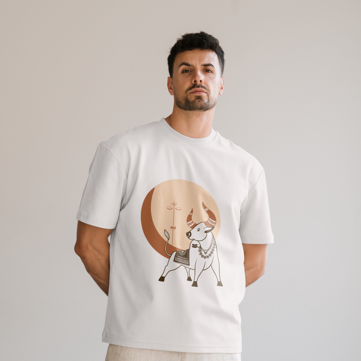 A man wearing a white crewneck t-shirt with a graphic print of a white nandi / bull in front of a large tan sun on the front, set against a plain light gray background.