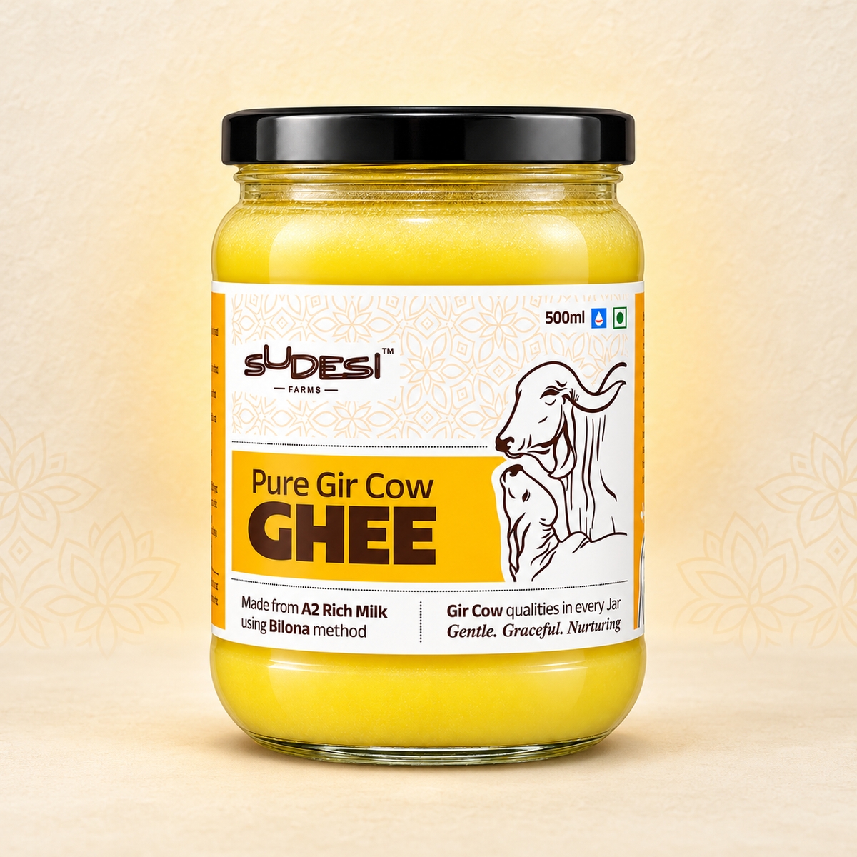 A 500ml glass jar of Sudesi Pure Gir Cow Ghee with a black lid and a white and yellow label, set against a light beige background with subtle floral patterns.
